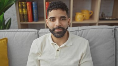 Samimi bir oturma odasında gri bir kanepede oturmuş gülümseyen sakallı genç bir İspanyol..