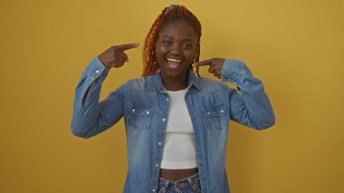 Kot ceketli neşeli bir Afrikalı kadın sarı arka planda gülümseyip yanaklarını işaret ediyor..
