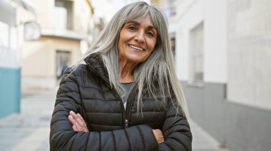 Şehir ortamında gri uzun saçlı, kollarını kavuşturmuş, sokak geçmişi olan orta yaşlı, kendine güvenen İspanyol bir kadın..