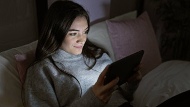 Latin kökenli genç bir kadın geceleri rahat bir yatak odasında dinleniyor, modern tabletini kullanıyor..