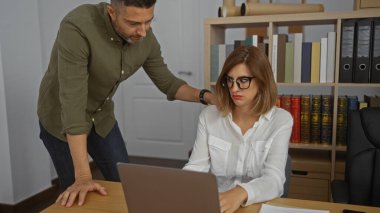 Kadın ve erkek, arka planda kitaplıklarla bir ofis ortamında birlikte çalışıyorlar. Dizüstü bilgisayarına odaklanmış kadının üzerine eğilen adamla birlikte.