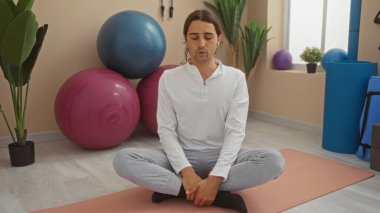 Arka planda yoga topları ve bitkilerle bir spor salonunda meditasyon yapan genç bir adam sakin ve odaklanmış bir çalışma atmosferini vurguluyor..