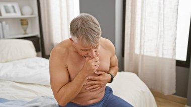 Senior man with grey hair shirtless sitting on a bed in pain holding chest implying potential heart attack indoors clipart