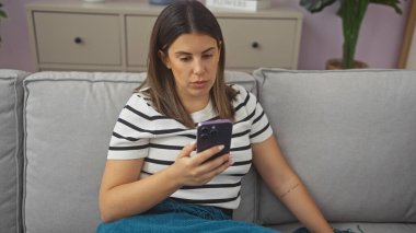 Odaklanmış genç bir kadın evdeki kanepede akıllı telefon kullanıyor, modern kapalı alan aktivitelerini tasvir ediyor..
