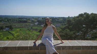 Beyaz elbiseli genç bir kadın kameraya gülümsüyor. Locorotondo, puglia, İtalya 'nın yemyeşil kırsalına bakan taş bir duvarda oturuyor..