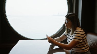 A brunette woman wearing glasses sits by a round window, looking at her phone on a cruise ship, capturing a sense of leisure travel. clipart