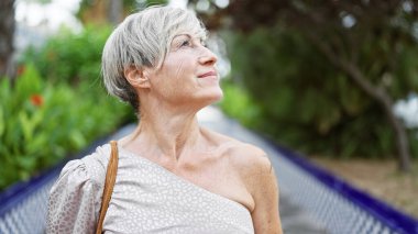 Kısa gri saçlı, düşünceli, olgun bir İspanyol kadın yemyeşil bir parkta huzurun tadını çıkarıyor..