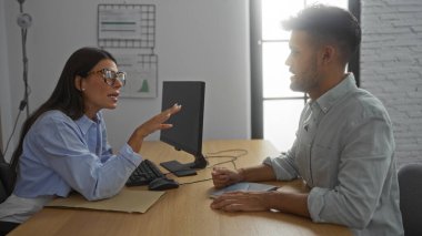 İşyerinde bir adamla konuşan bir kadın, bilgisayarla masanın karşısında oturuyor, parlak bir ofis ortamında iş konularını tartışıyor..