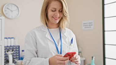 Laboratuvar önlüklü gülümseyen sarışın kadın klinikte akıllı telefon kullanıyor.
