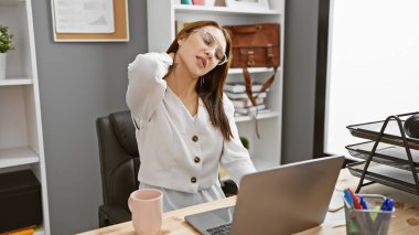Genç bir esmer kadın, modern bir ofis içinde dizüstü bilgisayarında çalışırken kendini rahatsız hissederek boynuna dokunuyor..