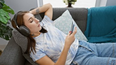 Genç bir kadın evdeki kanepede dinleniyor, akıllı telefonuyla kulaklıkla müzik dinliyor..