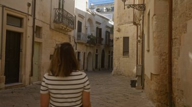 Lecce, Puglia, İtalya 'da büyüleyici dar bir caddeyi keşfeden genç bir kadın güneşli bir günde güzel tarihi binalar ve tuhaf balkonlarla çevrili..