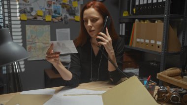 Odaklanmış kızıl saçlı bir kadın, soruşturma dosyaları ve şifreli bir haritayla karışmış bir dedektifin ofisinde telefonla konuşurken kanıtları analiz eder..