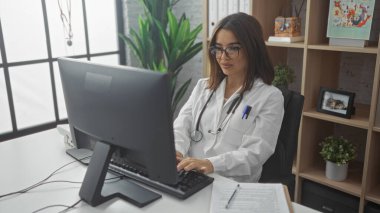 Kapalı bir klinikte çalışan çekici İspanyol kadın doktor, modern ofis ortamında bilgisayarına odaklanmış..