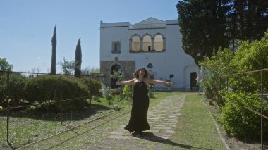 Siyah elbiseli güzel bir İspanyol kadın Puglia, İtalya 'daki tarihi bir villanın önünde neşeyle dans ediyor. Etrafı yemyeşil ve güneşli bir Avrupa manzarasıyla çevrili..