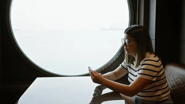 stock image A brunette woman wearing glasses sits by a round window, looking at her phone on a cruise ship, capturing a sense of leisure travel.