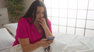 Kadın rahat bir yatak odasında pembe gömlek giyip akıllı telefon kullanırken gülümsüyor..