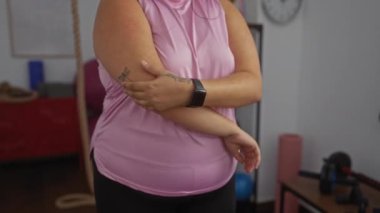 Ayrıca beden ölçülerinde bir kadın spor salonunda esniyor, pembe bir bluz giyiyor ve her iki kolunda da dövmeleri var ve arka planda spor malzemeleri var.