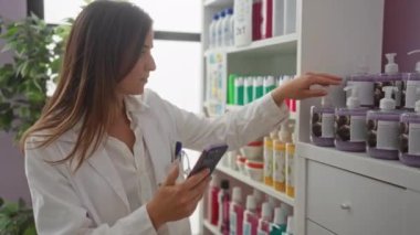 Eczanede beyaz önlük giyen ve akıllı telefon kullanan genç, sarışın bir kadın arka planda kapalı bir bitki bulunan raftaki çeşitli losyon şişelerini inceliyor..