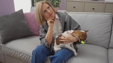 Olgun, sarışın, beyaz bir kadın oturma odasındaki kanepeye nazikçe oturur ve elinde bir topla küçük bir köpek tutar..