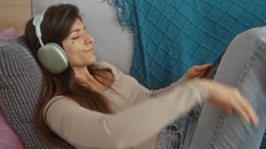 Genç bir İspanyol esmer kadın, oturma odasında kulaklıkla müzik dinlerken rahatça kanepede uzanıyor..