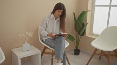 Genç bir İspanyol kadın modern bir bekleme odasında oturuyor, Minimalist bir iç mekan ortamında, panodaki belgelere dikkatlice bakıyor..
