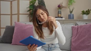 Gözlüklü genç beyaz bir kadın evinde rahat bir kanepede otururken tablet kullanıyor..
