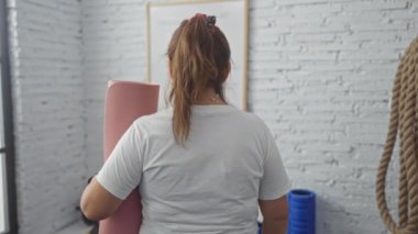 Kapalı spor salonunda pembe yoga minderi tutan olgun, İspanyol bir kadın. Arka planda spor aletleriyle kameraya gülümsüyor ve el sallıyor..