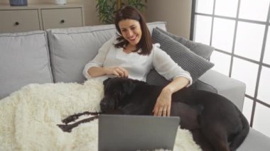 Şehir parkındaki genç bir İspanyol kadın video görüşmesi sırasında laptopuna gülümsüyor. Açık hava ortamında siyah labradoruyla rahat bir battaniyeye uzanıyor..