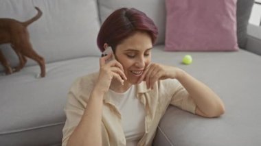 Kısa saçlı genç bir kadın akıllı telefonuyla sohbet ediyor. Oturma odasında rahat bir şekilde oturuyor. Arka planda neşeli bir köpek ve tenis topu var..