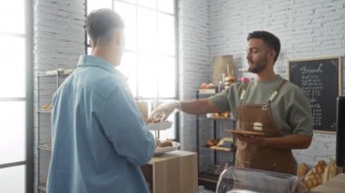Fırıncı, modern iç ve beyaz tuğla duvarlı, rafları ekmek ve hamur işleriyle dolu ve arka planda görünen fiyat menüsü olan kapalı fırındaki müşterilere hamur işleri sunuyor.
