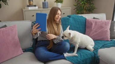 Genç, sarışın, beyaz, beyaz köpeğinin yanında kanepede otururken mavi tabletle bir video görüşmesi yapan beyaz bir kadın..