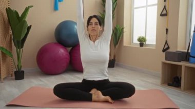 Spor salonunda yoga yapan İspanyol kadın, spor topu, bitki ve bantlar gibi spor malzemeleriyle çevrili minderin üzerinde geriniyor..