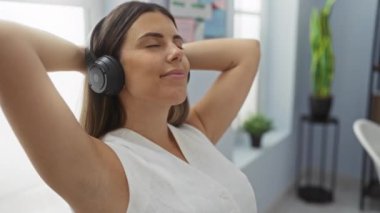Parlak bir ofiste kulaklıkla dinlenen ve yüzünde gülümsemeyle müziğin tadını çıkaran bir kadın.