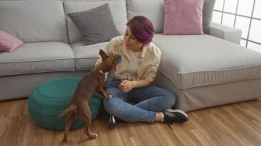 Kısa mor saçlı genç bir kadın, modern oturma odasının ahşap zemininde küçük kahverengi köpeğiyle oynuyor. Yeşil bir puf, gri bir kanepe ve renkli yastıkların yanında oturuyor..