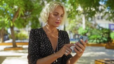 Şehir parkında, açık havada akıllı telefon kullanan bir kadın, ekrana odaklanmış, modern şehir arka planında, siyah benekli elbise giymiş, yeşil yapraklarla çevrili, doğal ışık, parlak bir gün.