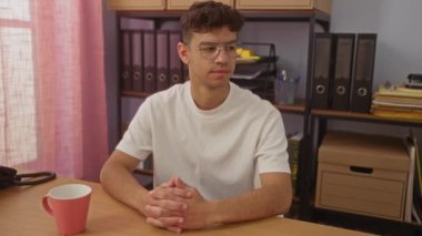 Ofiste oturan, gülümseyen, beyaz tişörtlü, pembe perdeli, dosyalı ve masasında kahve bardağı olan İspanyol asıllı genç bir adam..