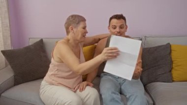 Anne ve yetişkin oğul birlikte belge okuyor ve oturma odasındaki kanepede sarılıyor. Aile sevgisini gösteriyor.