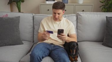 Evde oturan İspanyol asıllı genç adam oturma odasında bir kanepede oturuyor, elinde akıllı bir telefon ve bir kredi kartı, yanında da yanında bir köpekle online alışveriş yapıyor..