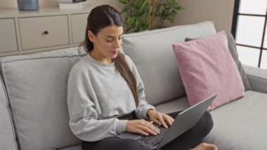 Oturma odasında oturan Latin kökenli genç bir kadın dizüstü bilgisayarını kapatıp kollarını kavuşturuyor. Gri bir kanepede oturmuş, rahat ve düşünceli bir atmosferi somutlaştırıyor..