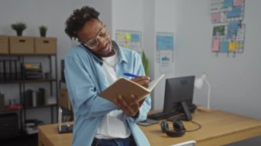 Bir ofis ortamında birden fazla iş yapan Afrikalı Amerikalı bir adam, modern bir iş yerinde rafları ve masasında bir bilgisayar olan bir not defterinde yazarken telefonla konuşuyor..