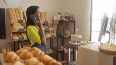 Ekmek ve hamur işleri dükkanının önünde telefonla konuşan bir kadın. Sarı tişört ve mavi önlük giyiyor. Parlak bir gülümsemesi var.