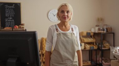 Önlüklü sarışın kadın gülümsüyor ve fırında ekmek ve hamur işleri sergileniyor.