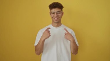 Beyaz tişört giyen genç İspanyol adam mutlu bir şekilde ayakta duruyor ve izole edilmiş sarı arka planda gülümsüyor.