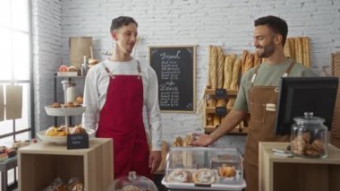 Erkekler fırında el sıkışıyor. Etrafı ekmek ve hamur işleriyle çevrili. Gülümsüyor ve önlük takıyor.