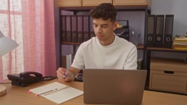 Modern bir ofiste dizüstü bilgisayarda çalışan, gözlük takan ve ofis malzemeleri, klasörler ve raflarla çevrili vazifelerine odaklanan genç İspanyol bir adam..