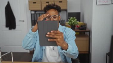 Genç, yakışıklı, Afro-Amerikalı, gözlüklü ve isim etiketli bir tablet kullanarak ofiste oturuyor. Raflı bir masada oturuyor ve arka planda bir bitki var..