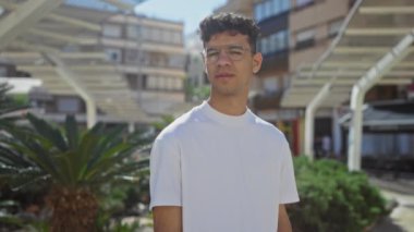 Güneşli bir günde, arka planda binalar olan gözlük takan İspanyol bir adam şehir parkında dikiliyor..