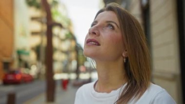 Güzel sarışın bir kadın gülümsüyor ve arka planda bulanık binalar olan bir şehir sokağında dikiliyor..