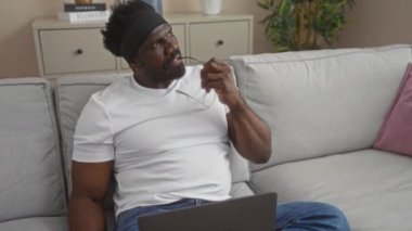 Düşünceli bir Afrikalı Amerikalı adam rahat bir oturma odasında dizüstü bilgisayarıyla bir koltukta oturur ve elinde gözlükleriyle oturup düşünür..